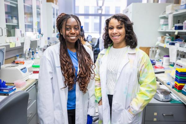Two female students of color smiling for photo wearing white lab coats in biomedical lab